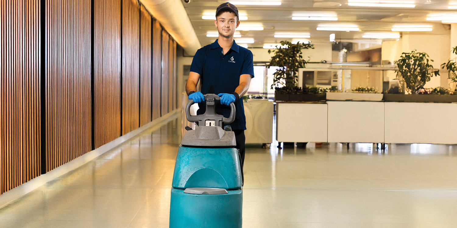 Employee operating a walk-behind floor cleaner wearing a dark shirt and looking straight ahead