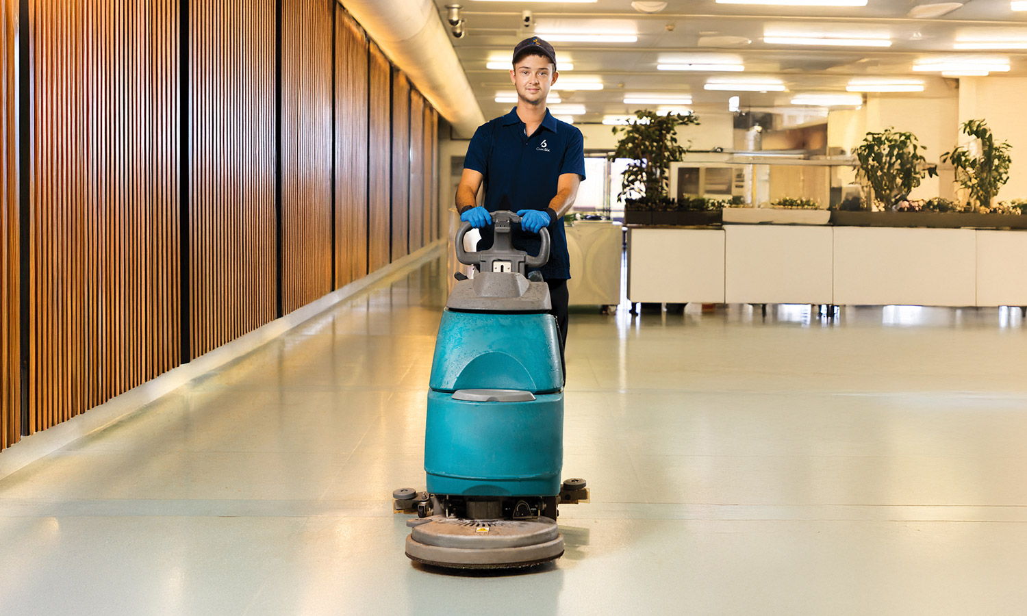 Employee operating a walk-behind floor cleaner wearing a dark shirt and looking straight ahead
