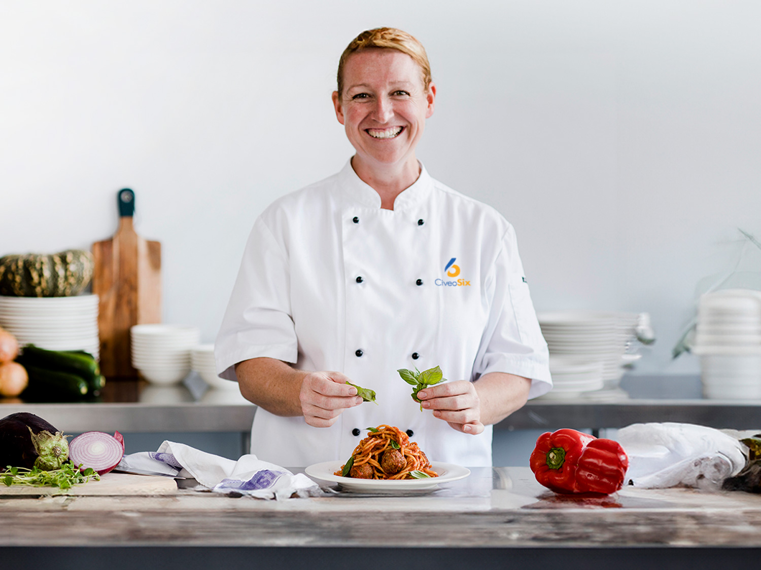 Chef in white uniform preparing a plate of pasta with fresh ingredients looking straight ahead